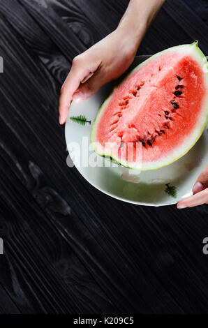 Scheibe Wassermelone auf weiße Platte in Frau Hände zum Essen bereit, Ansicht von oben mit verschwommenen schwarz Holz Tisch Hintergrund Stockfoto