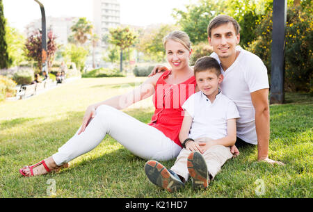 Lächelnde junge Familie von drei sitzen zusammen auf der grünen Wiese in der Stadt im Sommer Tag Stockfoto