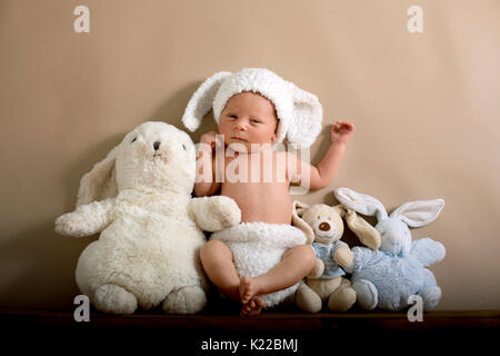 Neugeborenes Baby Boy trug ein braunes Kaninchen hat und Hosen aus Gewirken, schlafen auf einem Regal neben kleinen flauschigen Kaninchen. Im Studio Schuß auf eine cremige Hinterg Stockfoto