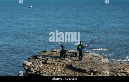 Zwei Angler mit Rod- und Linien-Seeangeln von Küstenklippen in Northumberland mit einem kleinen Boot und Seeanglern im Hintergrund Stockfoto