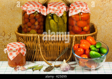 Eingemachtes Gemüse (Tomaten, Gurken, Tomaten) im Glas auf Weidenkorb, frisches Gemüse (Tomaten, Gurken) In Glasschale, Löffel mit Stockfoto