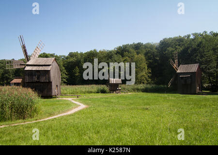 Drei alte hölzerne Windmühlen auf Gras in der Nähe einer Landstraße mit grünen Wald im Hintergrund Stockfoto