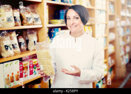 Lächelnd reife Frau Verkäufer in Uniform holding Getreide Produkte in den Händen in der Nähe von Regalen Stockfoto