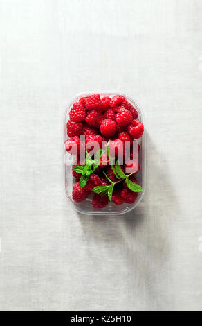 Frische Himbeeren in Kunststoffbox am weißen Stein Hintergrund. Stockfoto