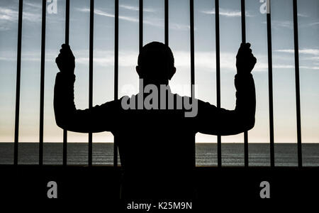 Silhouette des Menschen durch Stahl Bars mit Blick auf das Meer im Hintergrund. Depression, psychische Gesundheit, Asylbewerber, Einwanderung ... Konzept Bild. Stockfoto