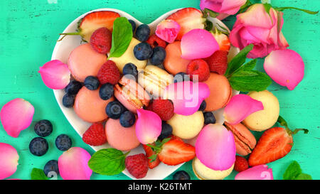 Petit Fours Präsentation der Macarons mit Beeren und Rosenblätter in Herzform auf rustikalen aqua grün Holz Tisch, bis zu schließen. Stockfoto