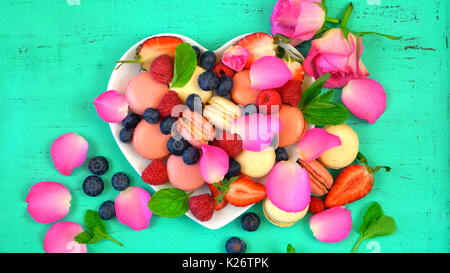 Petit Fours Präsentation der Macarons mit Beeren und Rosenblätter in Herzform auf rustikalen aqua grün Holz Tisch Stockfoto