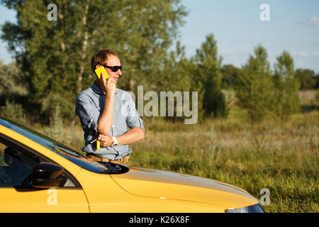 Ein Mann ist über sein Mobiltelefon. Er steht neben dem gelben Auto. Kommunikation in der Reise. Stockfoto