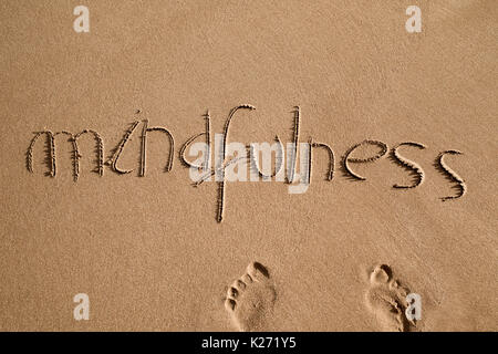 High-Angle Shot des Wortes Achtsamkeit in den Sand des Strandes geschrieben und ein paar menschliche Fußabdrücke Stockfoto