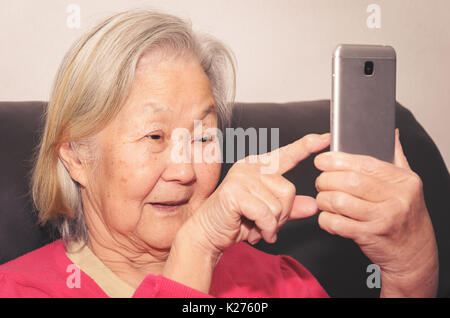 Alte Frau auf einem Sofa zu Hause Holding ein Smartphone sitzt und den Bildschirm berühren mit dem Finger. Stockfoto