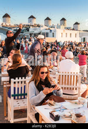 Touristen in einem Café Waterfront mit dem Kato Mili Windmühlen hinter Ihnen, Mykonos, Griechenland sitzen. Stockfoto