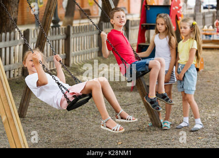Blick auf fröhliche ausgelassene Kinder zusammen Schwingen auf Kinderspielplatz in der Stadt Stockfoto