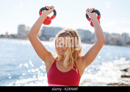 Glückliche junge blonde Frau Workout mit kettlebell Outdoor an der Küste Stockfoto