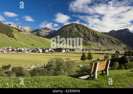 Große Stadt in der Ferne und Holzbank Nahaufnahme mit hohen grünen Bergen im Hintergrund und bewölktem Himmel Stockfoto