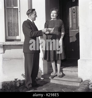 1964, historische, Bild zeigt einen elegant gekleideten Gentleman präsentiert einen Check an einen überrascht und ein wenig schockiert housewfie noch immer in ihren Hausschuhen vor der Tür am Eingang zu Ihrem Haus, England, UK. Stockfoto