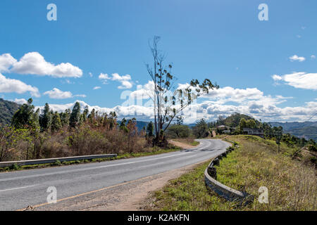 Leere ländlichen geschwungene Asphalt Land straße durch trockenen Winter vegetation Landschaft gegen den blauen bewölkten Himmel in KwaZulu-Natal in Südafrika Stockfoto