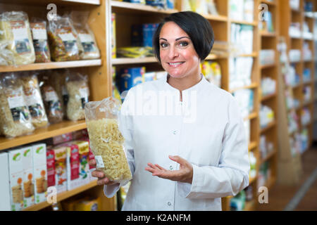 Lächelnd reife Frau Verkäufer in Uniform holding Getreide Produkte in den Händen in der Nähe von Regalen Stockfoto