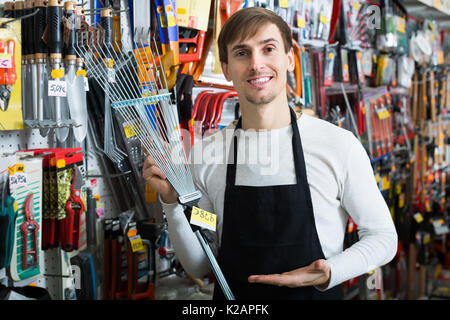 Positive junge Verkäufer in Schürze mit Blatt rake im Supermarkt Stockfoto