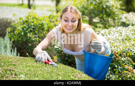 Glücklich lächelnde junge blonde Frau, die Pflege des grünen Büsche in den Garten im Sommer Tag Stockfoto