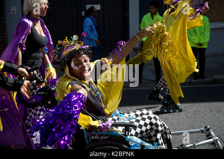 Notting Hill Carnival August 2017. Ein Karneval Nachtschwärmer Tänze in ihrem Rollstuhl, neben einer Gruppe von Walze bladers. Stockfoto