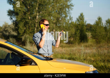 Ein Mann ist über sein Mobiltelefon. Er steht neben dem gelben Auto und hält den Zündschlüssel in seinen Händen. Kommunikation in der Reise. Stockfoto