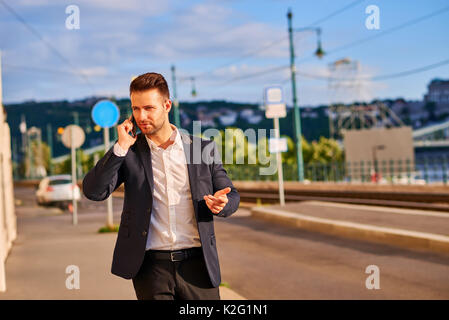 Ein hübscher junger Unternehmer gehen auf die Straße am Fluss, der gerade sein Telefon Stockfoto