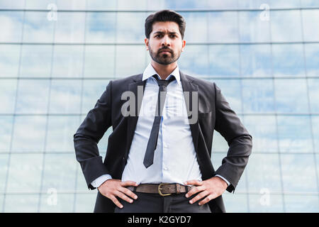 Arabisch ernsthafte Geschäftsmann oder Arbeitnehmer im schwarzen Anzug mit Krawatte und Hemd mit Bart stand vor einem Bürogebäude auf grünem Gras im Sommer Tag. Stockfoto