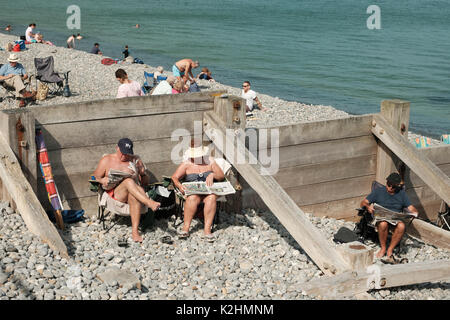 Paar mittleren Alters lesen Zeitungen in Liegestühlen auf einem Kieselstrand in Norfolk, Großbritannien, an einem heißen Wochenende Stockfoto