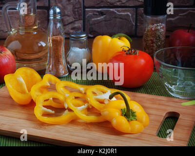 Große gelbe saftig geschnitten Pfeffer Ringe auf einer hölzernen Schneidebrett auf einem Tisch mit roten Tomaten, Äpfel, Pflanzenöl, Salz, Gewürze Stockfoto