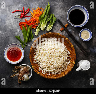 Raw Udon Nudeln in Bambus Korb und Zutaten zum Kochen asiatische Lebensmittel mit Tiger Garnelen, Grüns, Gemüse, Gewürze auf dunklem Hintergrund Stockfoto