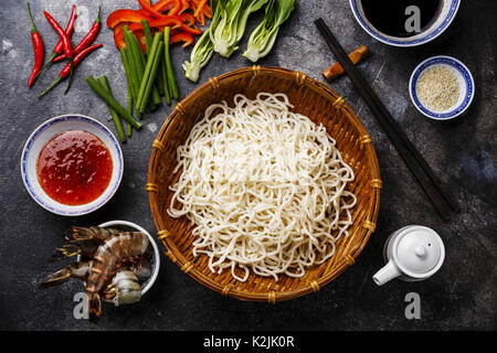 Raw Udon Nudeln in Bambus Korb und Zutaten zum Kochen asiatische Lebensmittel mit Tiger Garnelen, Grüns, Gemüse, Gewürze auf dunklem Hintergrund Stockfoto