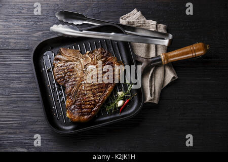 Gegrillte chemische Alterung T-Bone Steak im Grill Bügeleisen pan und Zange auf schwarz verbrannten Holz- Hintergrund Stockfoto