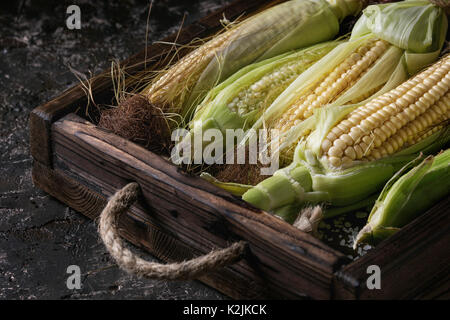 Junge Roh ungekocht Maiskolben in Blätter in Holz- Schließen. Über Dunkelbraun konkrete Textur Hintergrund. Stockfoto