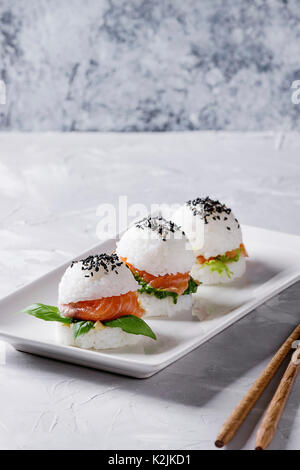 Mini Reis sushi Burger mit geräuchertem Lachs, grüner Salat und Saucen, schwarzer Sesam auf weiße quadratische Platte mit hölzernen Eßstäbchen über Graues konkret serviert. Stockfoto