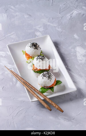 Mini Reis sushi Burger mit geräuchertem Lachs, grüner Salat und Saucen, schwarzer Sesam auf weiße quadratische Platte mit hölzernen Eßstäbchen über Graues konkret serviert. Stockfoto