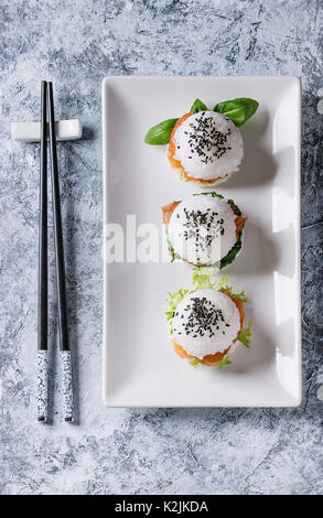 Mini Reis sushi Burger mit geräuchertem Lachs, grüner Salat und Saucen, schwarzer Sesam auf weiße quadratische Platte mit Stäbchen über grauer Beton Hinterg serviert. Stockfoto