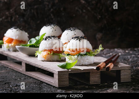 Mini Reis sushi Burger mit geräuchertem Lachs, grüner Salat und Saucen, schwarzer Sesam auf Holzpalette Fach mit Stäbchen über Dunkelbraun konkrete serviert b Stockfoto
