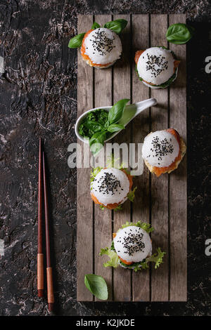 Mini Reis sushi Burger mit geräuchertem Lachs, grüner Salat und Saucen, schwarzer Sesam auf Holzpalette Fach mit Stäbchen über Dunkelbraun konkrete serviert b Stockfoto