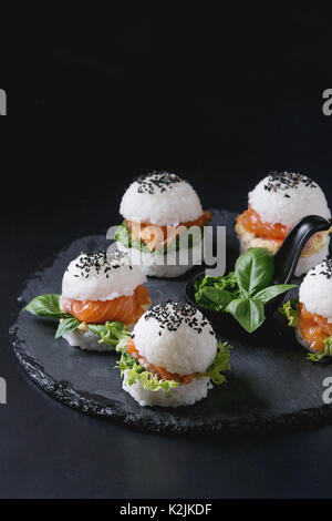 Mini Reis sushi Burger mit geräuchertem Lachs, grüner Salat und Saucen, schwarzer Sesam auf schiefer Brett über schwarzen Hintergrund serviert. Moderne gesunde foo Stockfoto