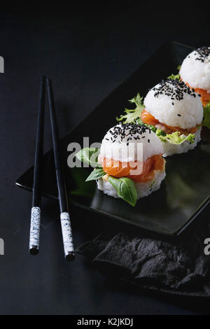 Mini Reis sushi Burger mit geräuchertem Lachs, grüner Salat und Saucen, schwarzer Sesam auf Schwarze quadratische Platte mit Stäbchen und textile Serviette serviert über Stockfoto