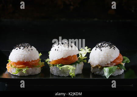 Mini Reis sushi Burger mit geräuchertem Lachs, grüner Salat und Saucen, schwarzer Sesam auf Schwarze quadratische Platte mit textilen Serviette serviert über schwarzen backgroun Stockfoto