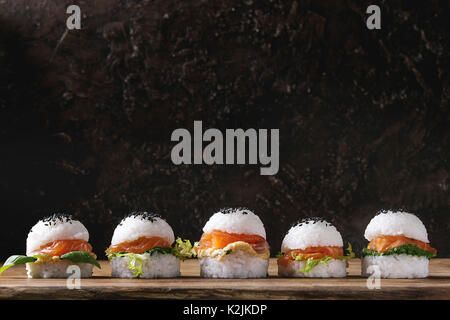 Mini Reis sushi Burger mit geräuchertem Lachs, grüner Salat und Saucen, schwarzer Sesam mit Stäbchen aus Holz Über Holz Hintergrund serviert. Moderne gesunde fo Stockfoto