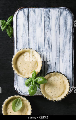 Teig zum backen Quiche Tarte in kleinen backen Form bereit für Backen mit frischem Basilikum auf weißem Holz das Brett über Schwarze verbrannte Holz- Hintergrund. Fla Stockfoto