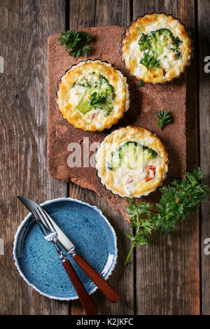 Gebackene Hausgemachte Quiche pie in Mini Metall Formen mit frischen Grüns, Teller und Besteck auf Terrakotta Board auf Old Plank Holz- Hintergrund serviert. Flach Stockfoto