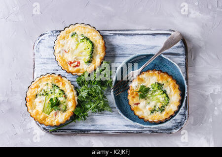 Gebackene hausgemachte Gemüse broccoli Quiche pie in Mini Metall Formen mit frischen Grüns, Teller, Gabel auf Weiß, das Board auf grauem Beton backgro Stockfoto