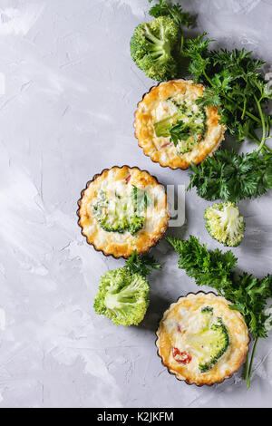 Gebackene hausgemachte Gemüse broccoli Quiche pie in Mini Metall Formen mit frischen Grüns auf grauem Beton Hintergrund serviert. Flach mit kopieren. Bereit Stockfoto