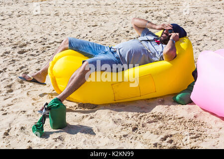 Bournemouth, Dorset, Großbritannien. September 2017. UK Wetter: Schöner warmer sonniger Tag am Bournemouth Strand an der Südküste. Mann entspannt sich auf gelben aufblasbaren Couch Liege sprechen auf Handy. Quelle: Carolyn Jenkins/Alamy Live News Stockfoto