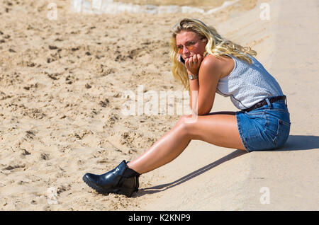 Bournemouth, Dorset, Großbritannien. September 2017. UK Wetter: Schöner warmer sonniger Tag am Bournemouth Strand - Besucher kommen früh dorthin, um einen guten Platz zu bekommen. Attraktive junge Frau mit langen blonden Haaren tragen Mini-Jeansrock sitzen am Strand halten Handy an ihr Ohr. Quelle: Carolyn Jenkins/Alamy Live News Stockfoto