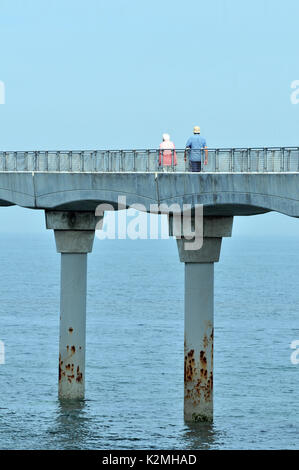 Ein pensionierter ältere ältere Paar entlang eine Brücke weg Hand in Hand über das Meer an der Küste im Urlaub bleiben aktiv in den Ruhestand Stockfoto