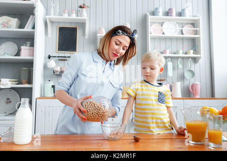 Mutter und Sohn lächelnd, während sie das Frühstück in der Küche. Mom gießt Milch und Cornflakes in Glas Stockfoto